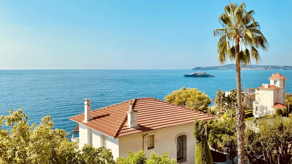 Casa con techo de tejas rojas y chimeneas entre árboles y palmeras, frente al mar