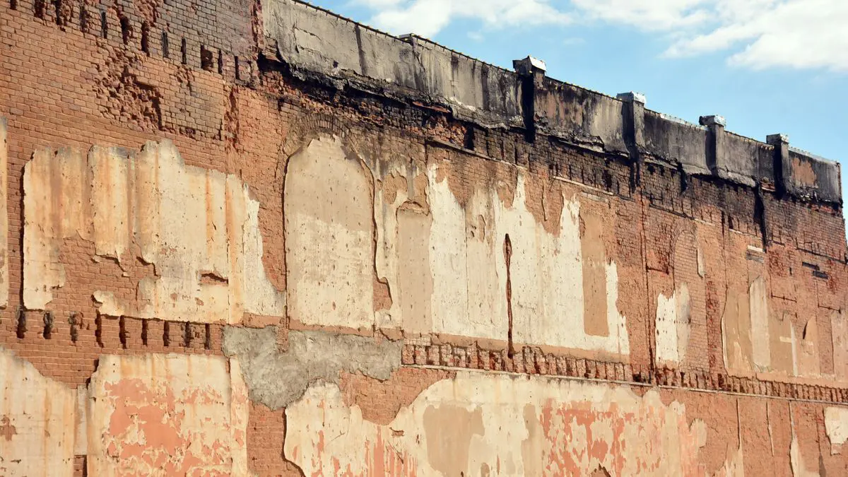 Muro alto de ladrillo con el revoque desprendido en parches y el remate deteriorado