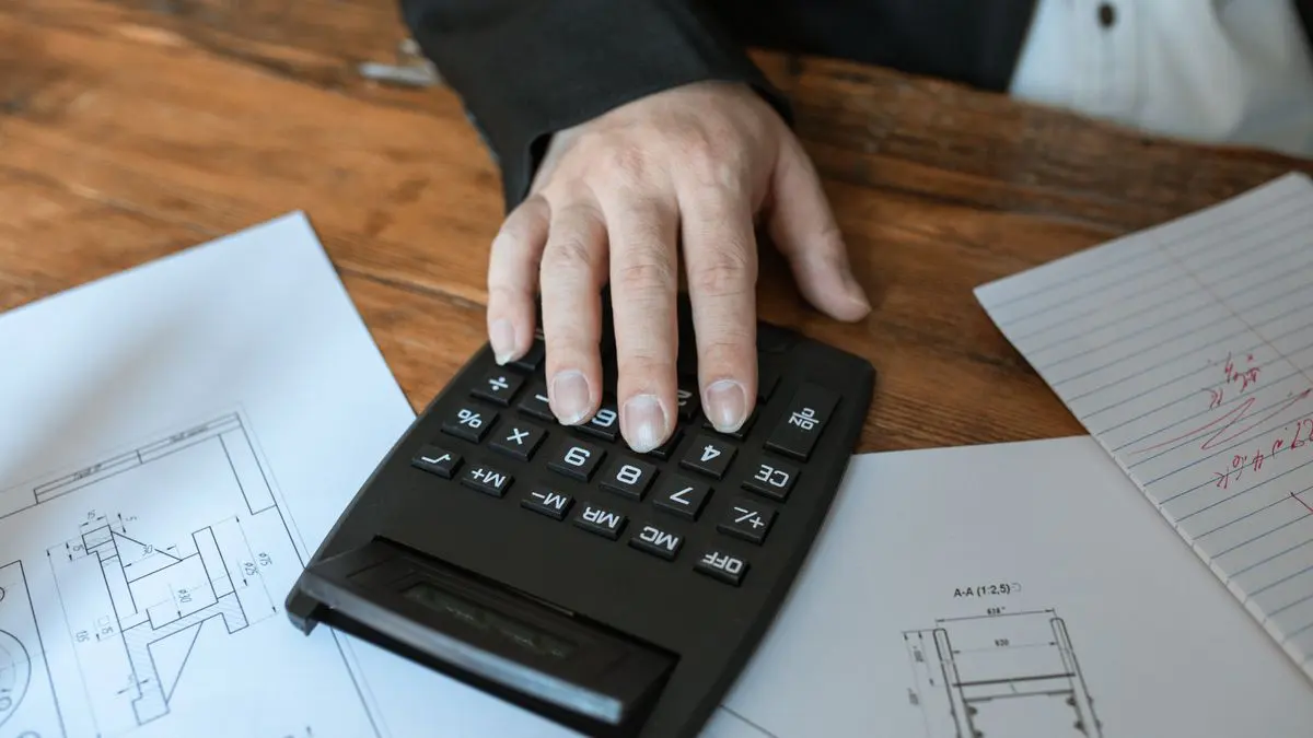 Mano usando una calculadora sobre planos y una hoja con anotaciones